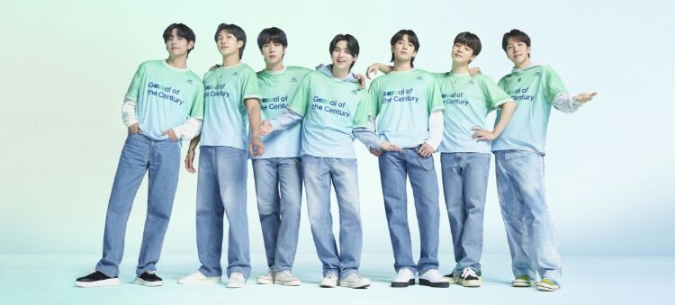 All 7 BTS members huddled together. They are all wearing blue jeans and a Team Century shirt with Hyundai logo and Goal of the Century emblazoned in dark blue across the front. RM, J-Hope and Suga are holding their arms in the air in celebration.