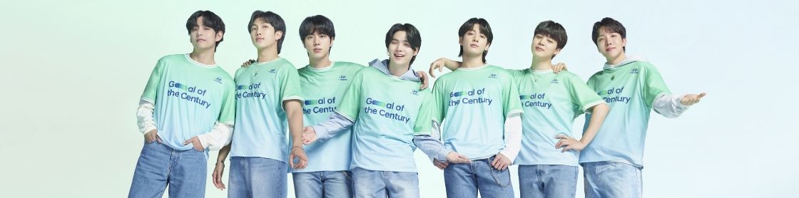 All 7 BTS members huddled together. They are all wearing blue jeans and a Team Century shirt with Hyundai logo and Goal of the Century emblazoned in dark blue across the front. RM, J-Hope and Suga are holding their arms in the air in celebration. 
