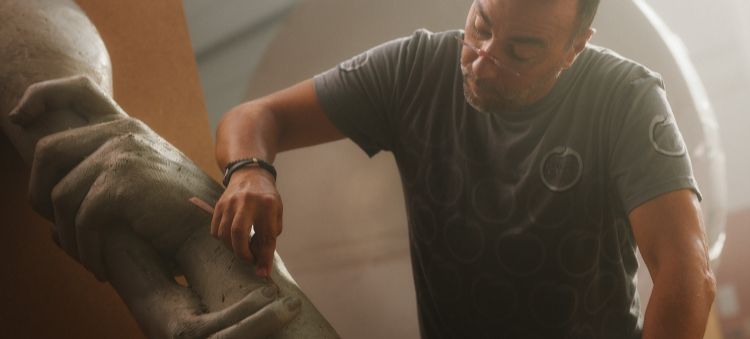Team Century member Lorenzo Quinn wearing glasses and a grey t-shirt while using a tool to shape the finger of an oversized clay sculpture of two hands.