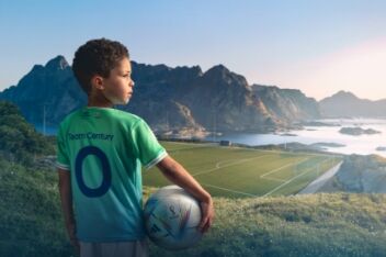 A young boy with dark hair wearing a Team Century jersey and looking out over a series of mountains and a lake from the top of a mountain.
