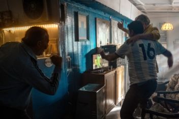 Two men in football jerseys hugging and celebrating while watching football on the television in a lounge. And a third man celebrates from the kitchen