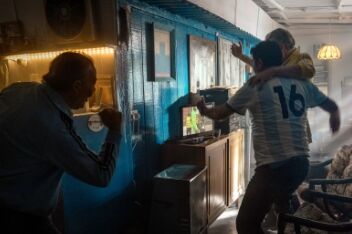 Two men in football jerseys hugging and celebrating while watching football on the television in a lounge. And a third man celebrates from the kitchen.
