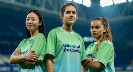 Three women standing in the middle of a stadium wearing green jerseys which say “Goal of the Century” on the front. 