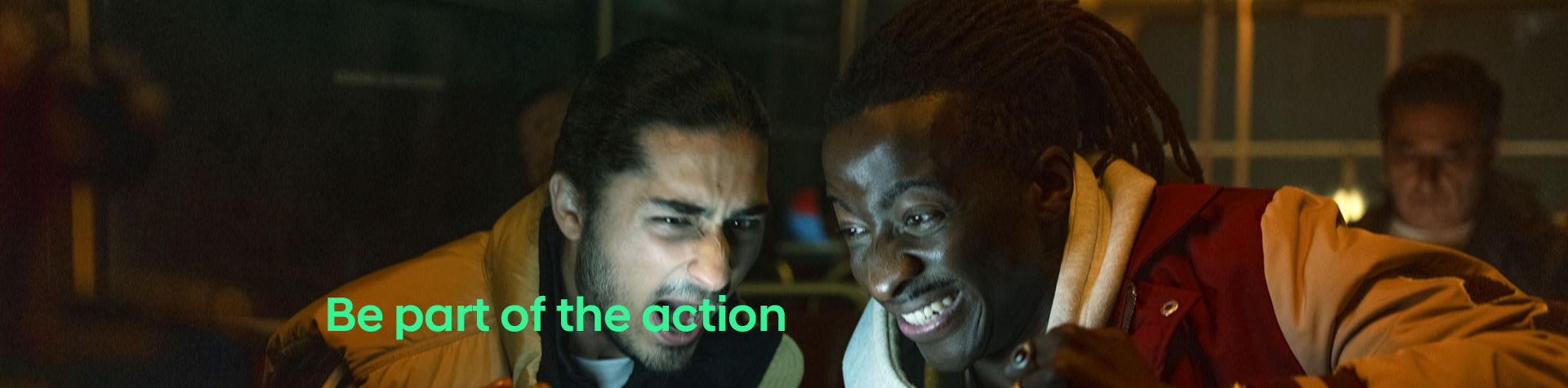 A close-up of the faces of two men watching a game of football and cheering. “Be part of the action” is written in green across the image.