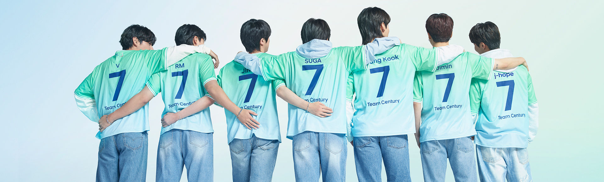 All 7 BTS members standing in a line facing backwards. Each member is wearing blue jeans with a green and gray Team Century shirt on with the number 7 emblazoned on the back in dark blue.