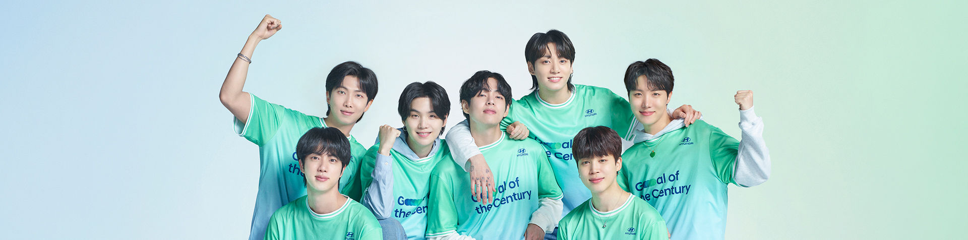 All 7 BTS members standing in a line facing forward. Each member has a green and gray Team Century shirt on with the Hyundai logo and Goal of the Century emblazoned in dark blue across the front. 