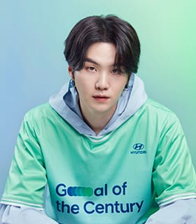 BTS member Suga wearing a green Team Century shirt with a gray hoodie underneath.