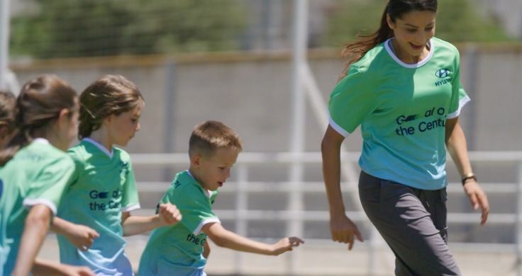 Team Century member Nadia Nadim and three kids are playing football. They are all wearing a green and gray Team Century shirt with Hyundai logo and Goal of the Century emblazoned in dark blue across the front.
