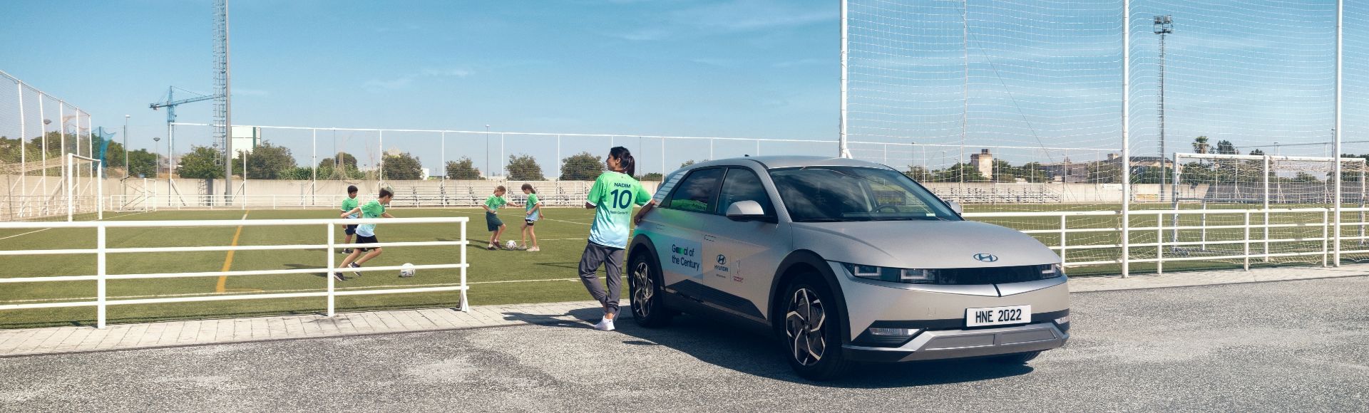 스페인 세비야에 있는 풋볼 마스 재단을 방문한 축구선수 나디아 나딤이 푸른색 Team Century 유니폼을 입고 IONIQ 5에 기대어 축구장에 있는 어린이들을 바라보는 모습입니다. 