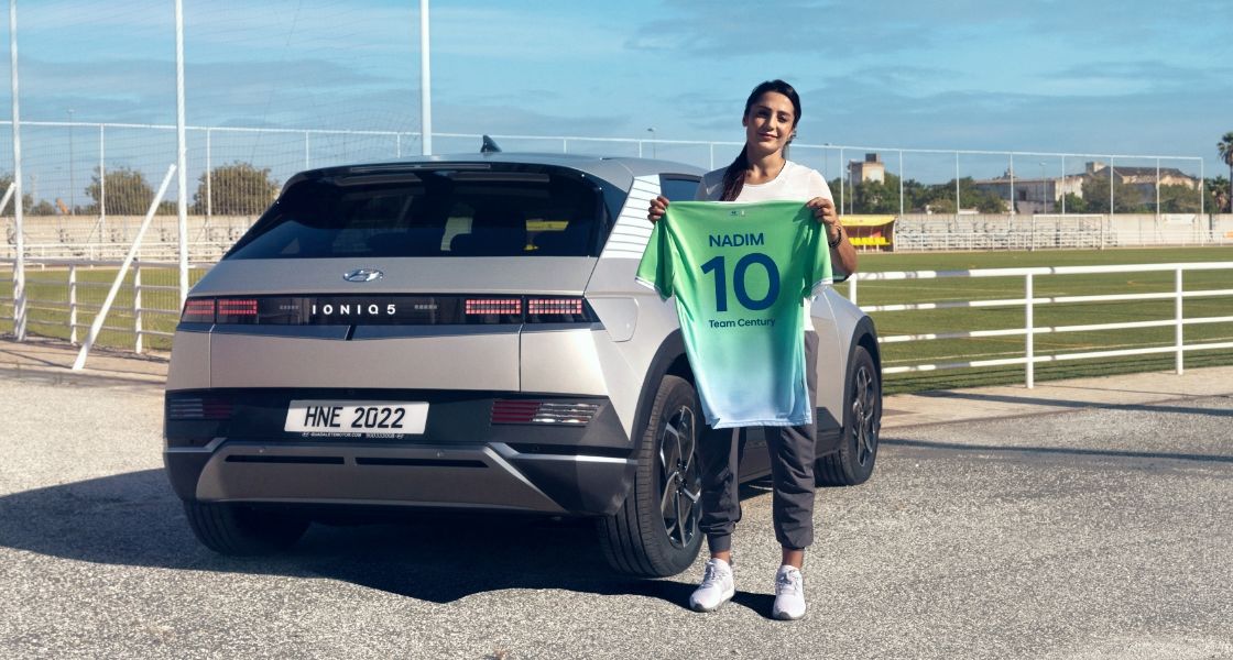 Footballer and Team Century member Nadia Nadim standing in front of a silver-coloured Hyundai IONIQ 5 next to a football field holding her green and blue Team Century shirt showing her name and the number 10.