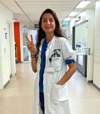 Denmark footballer and surgeon Nadia Nadim standing in a white hallway of a hospital in her white doctor’s coat smiling into the camera and giving the peace sign with her right hand.