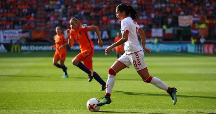 Female football player Nadia Nadim in red and white Danish national team jersey dribbling soccer ball with two female soccer players in orange jerseys running toward her.