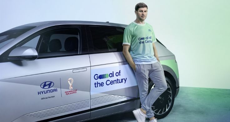 앞문에는 '현대'와 'FIFA 월드컵 2022' 스티커가, 뒷문에는 '세기의 골' 스티커가 붙은 실버 색상의 현대 아이오닉5에 스티븐 제라드가 파란색과 녹색의 '세기의 골' 티셔츠를 입고 기대어 서 있다.