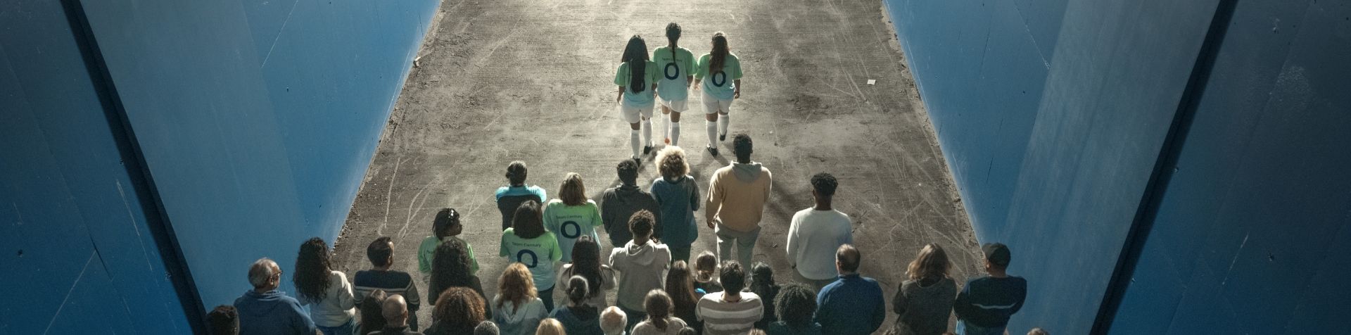 경기장으로 이어지는 복도를 걷는 한 무리의 축구 팬의 뒷모습을 위에서 바라본 모습. 긴 머리를 한 3명의 여성 축구 선수가 등에 파란색 숫자 0이 새겨진 녹색 Team Century 셔츠와 흰색 반바지, 축구 양말, 신발을 신고 몇 걸음씩 그룹을 이끌고 있습니다.
