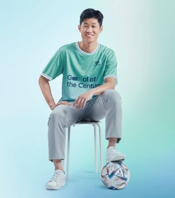 Team Century male footballer Jisung Park leaning forward while sitting in a chair with his left foot perched on a soccer ball and waering a light green Goal of the Century shirt.
