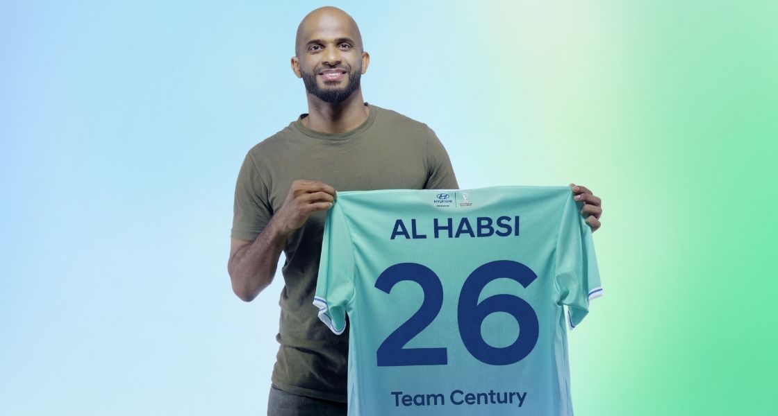 Torso view of new Team Century member Ali Al-Habsi smiling into the camera and holding up a green Team Century shirt with the  number 26 and Team Century emblazoned in blue across the back.
