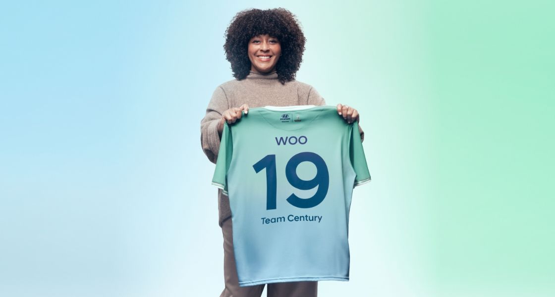 Nicky Woo standing and holding her Team Century jersey. She is wearing a brown jumper and brown trousers and smiling.