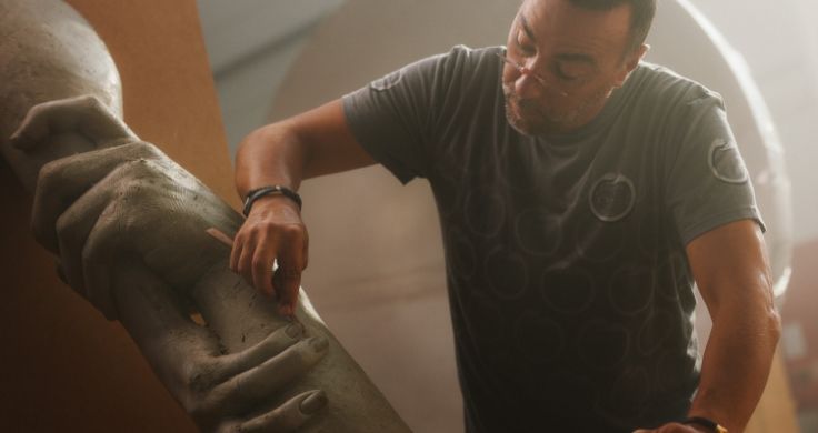 Team Century member Lorenzo Quinn wearing glasses and a grey t-shirt while using a tool to shape the finger of an oversized clay sculpture of two hands.