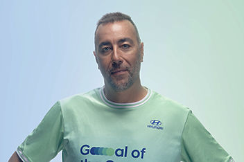 Portrait of world-renowned sculptor and new Team Century member Lorenzo Quinn smiling into the camera in front of a blue and green background.