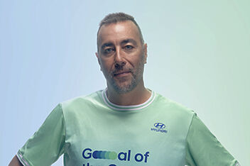 Portrait of world-renowned sculptor and new Team Century member Lorenzo Quinn smiling into the camera in front of a blue and green background.