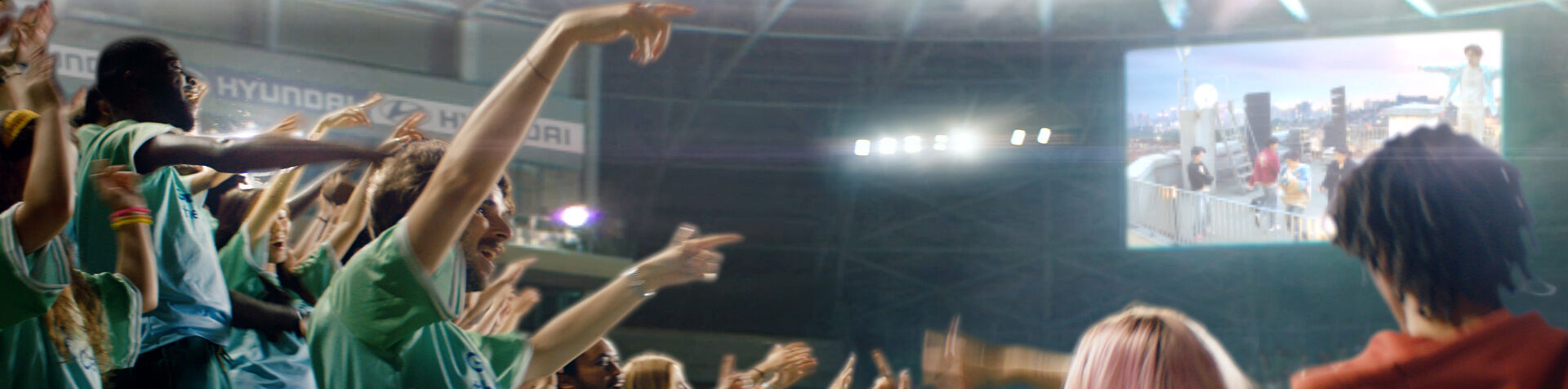 Crowd members all standing and cheering inside a football stadium watching a big screen on the other side. 