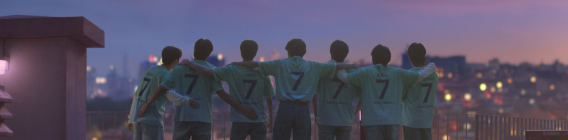 All 7 members of BTS standing on a rooftop at dusk with their backs to the camera. They are all wearing their Goal of the Century jerseys with the number 7 and BTS written on the back. 