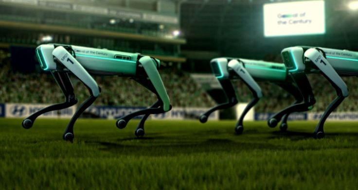 축구장에 나란히 서 있는 두 개의 로봇개 Spot 뒤에 세기의 골이 적힌 하얀 스크린이 있다.