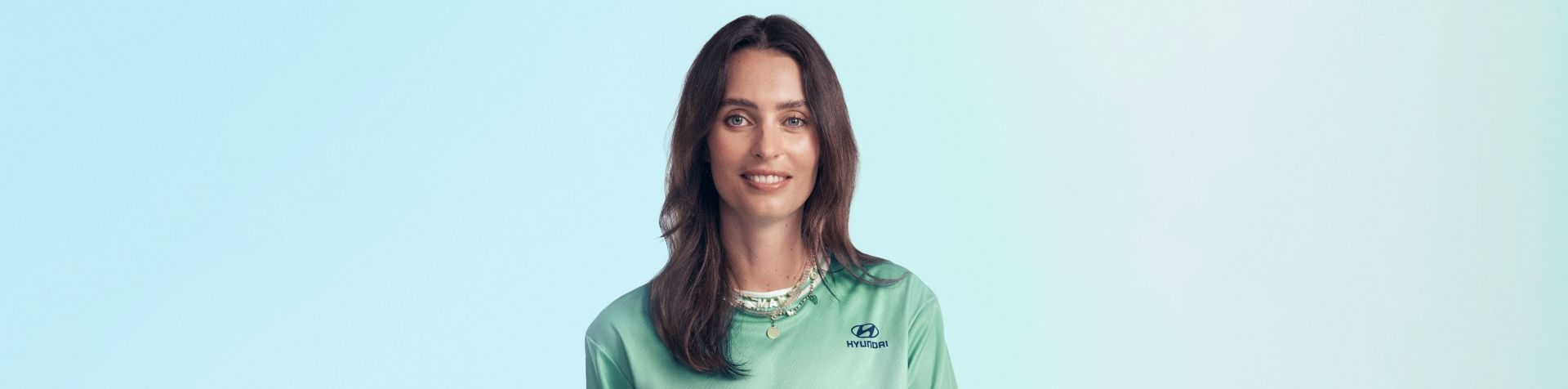 A close up of Ella Mills’ face. She is standing in front of a blue and green background and is wearing her green Team Century jersey and a couple of necklaces. She has long brown hair and blue eyes.