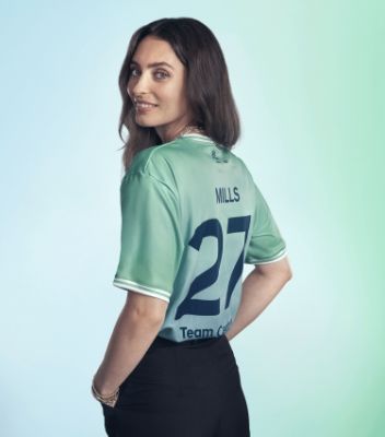Ella Mills standing with her back to the camera. She is standing in front of a light background, she she is wearing a green Team Century jersey with Mills, 27 and Team Century written on the back. 