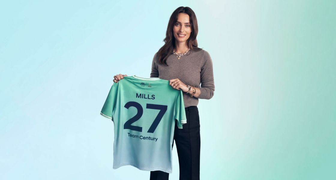엘라 밀스가 Team Century 유니폼을 손에 들고 카메라를 마주 보고 서있습니다. 그녀는 회색 스웨터와 검은색 바지를 입고 있습니다. 초록색과 파란색이 어우러진 그녀의 Team Century 유니폼 뒷면에는 ‘Mills’, ‘27’, ‘Team Century’ 문구가 쓰여 있습니다.