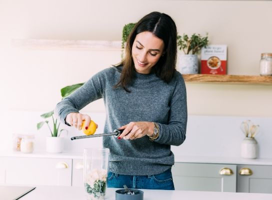Ella Mills standing behind a kitchen counter grating lemon zest into a cup filled with other ingredients. She is wearing a dark blue jumper and blue jeans. 
