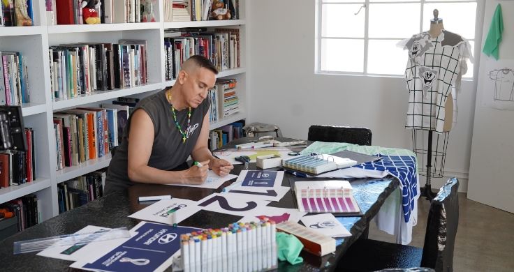 Jeremy Scott sitting at a desk in his studio. There are bookshelves behind him and lots of papers scattered around this desk. He is working on the Team Century shirt.
