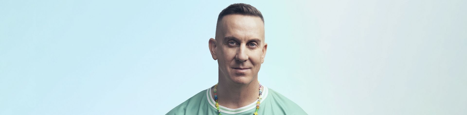 A close-up of Jeremy Scott’s head and shoulders. He has dark hair and is wearing a colorful necklace and the green Team Century shirt.