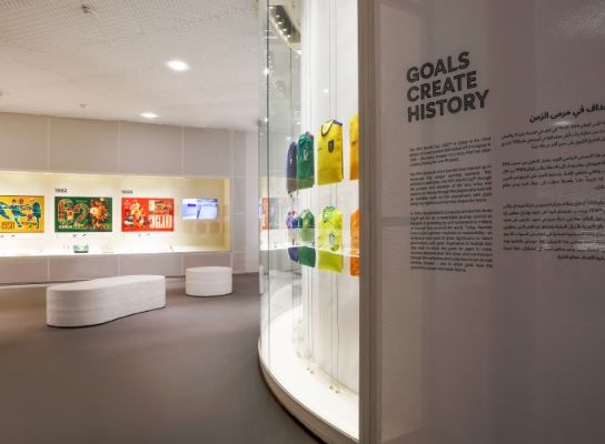 Inside the FIFA Museum building. There is an exhibition entitled Goals Create History and there are artwork and colorful football jerseys on display on the walls and in display cabinets.