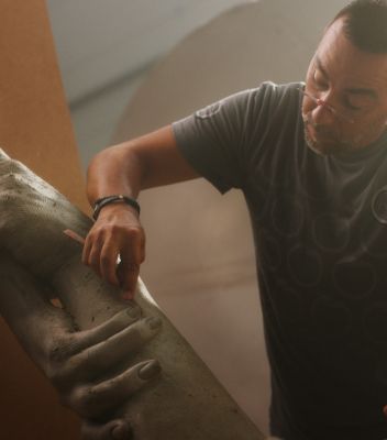 자신의 조각품 'The Greatest Goal'을 작업 중인 조각가 로렌초 퀸은 안경과 회색 티셔츠를 입고 조각품의 한쪽 팔을 다듬고 있습니다.