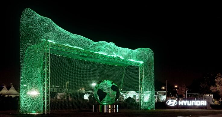 A large goal lit up in green at night time with a statue of a globe in the middle and the Hyundai logo lit up in white to the side.