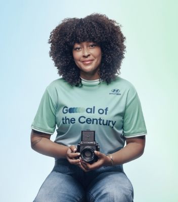 Nicky Quamina-Woo sitting down with her black camera in her lap. She is wearing the green and blue Team Century jersey and jeans and she has dark curly hair.