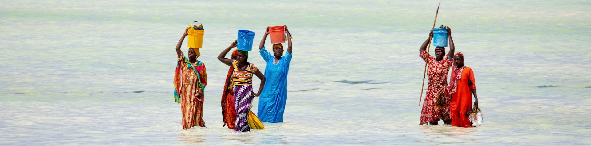 밝은색의 아프리카 전통 드레스와 스커트를 입은 세네갈 여성 여섯 명이 머리에 색색의 양동이를 이고 무릎 높이까지 오는 물속을 걷고 있습니다.